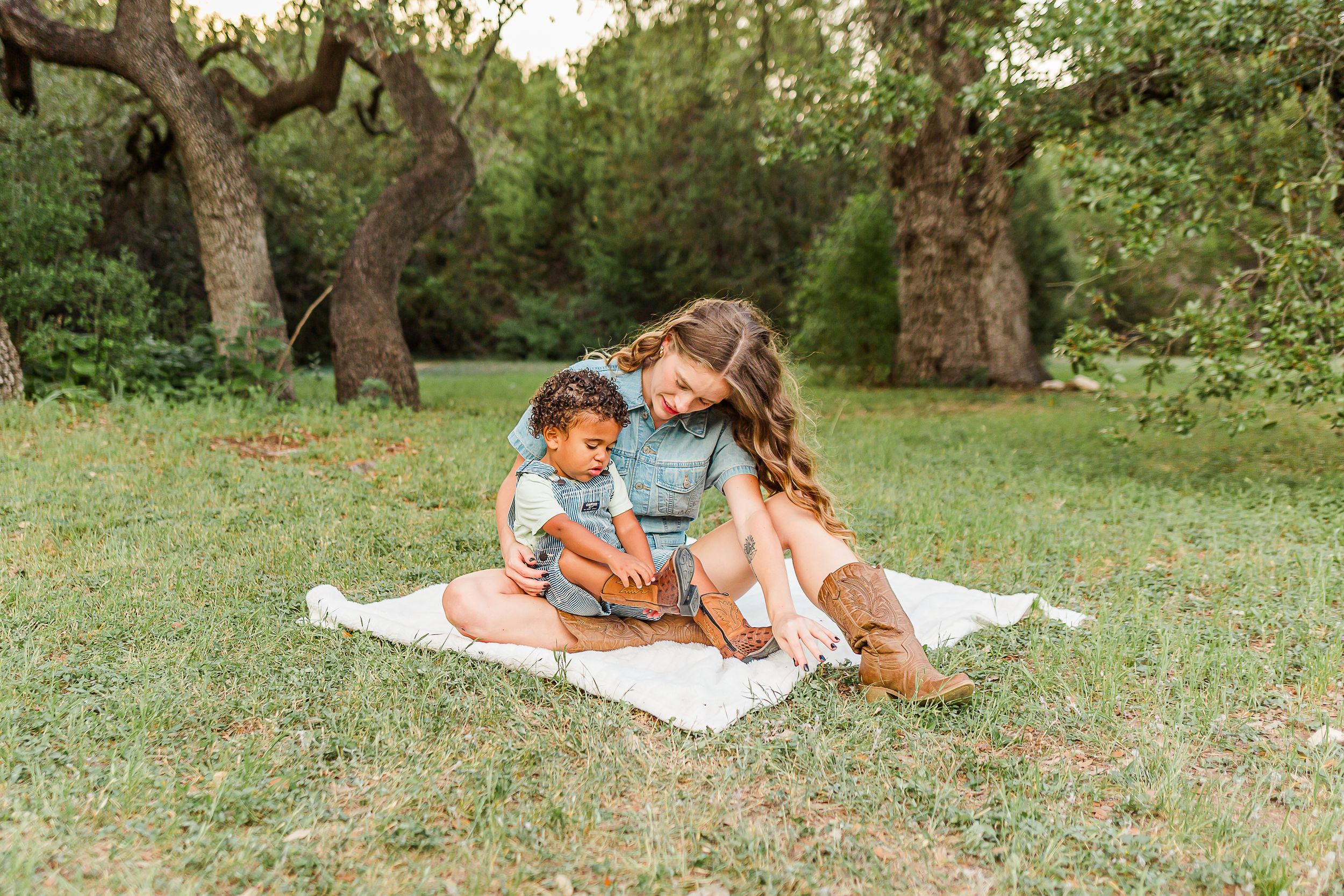 Two people sit together on a white blanket in a grassy field surrounded by old olive trees.
