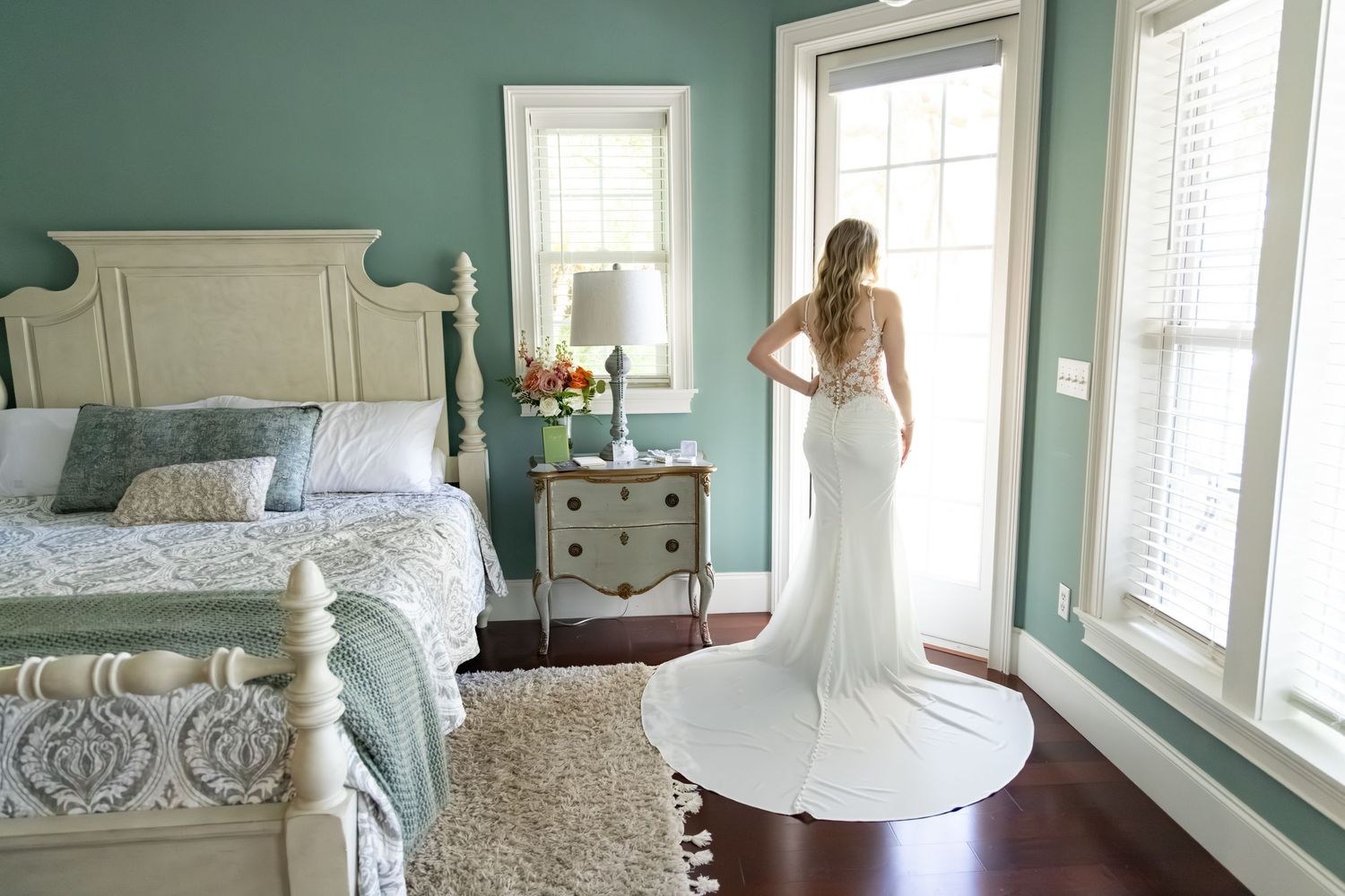 Bride in elegant white wedding gown with train standing by sunlit windows in a teal bedroom.
