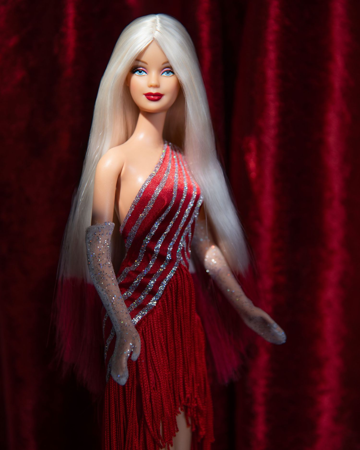 Barbie doll in red fringe sequin dress with long white hair posed against red velvet backdrop.