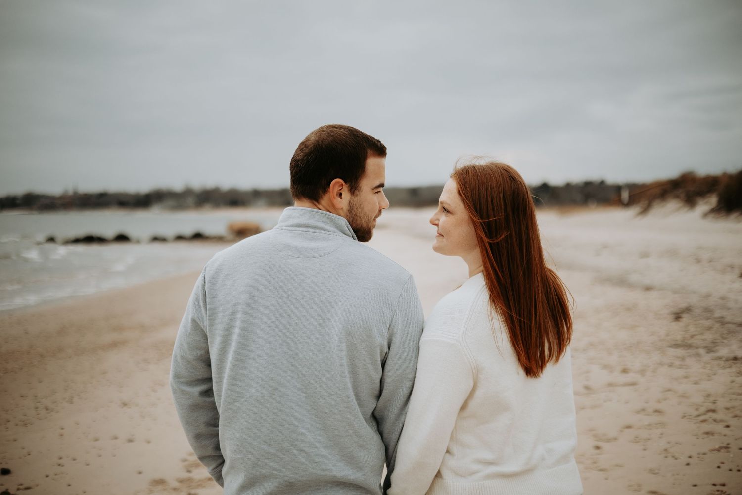 A couple walks along a misty beach shoreline in winter wearing coats and holding hands.