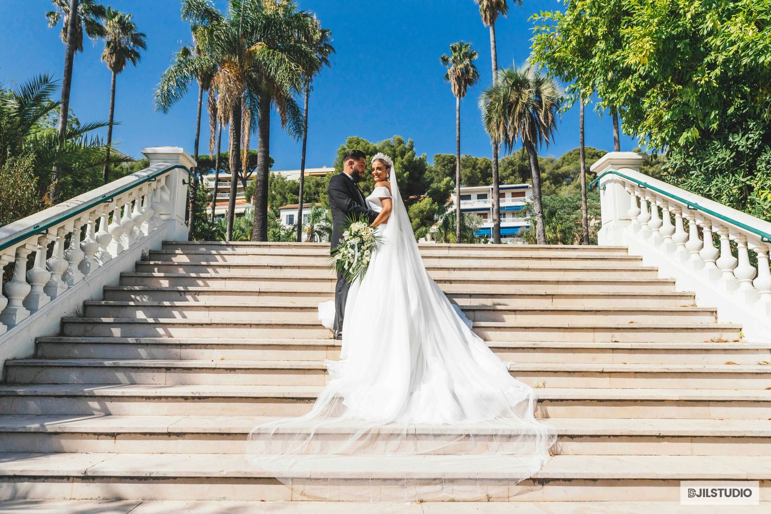 A bride in a white wedding dress gracefully ascends an elegant outdoor staircase surrounded by palm trees and greenery.