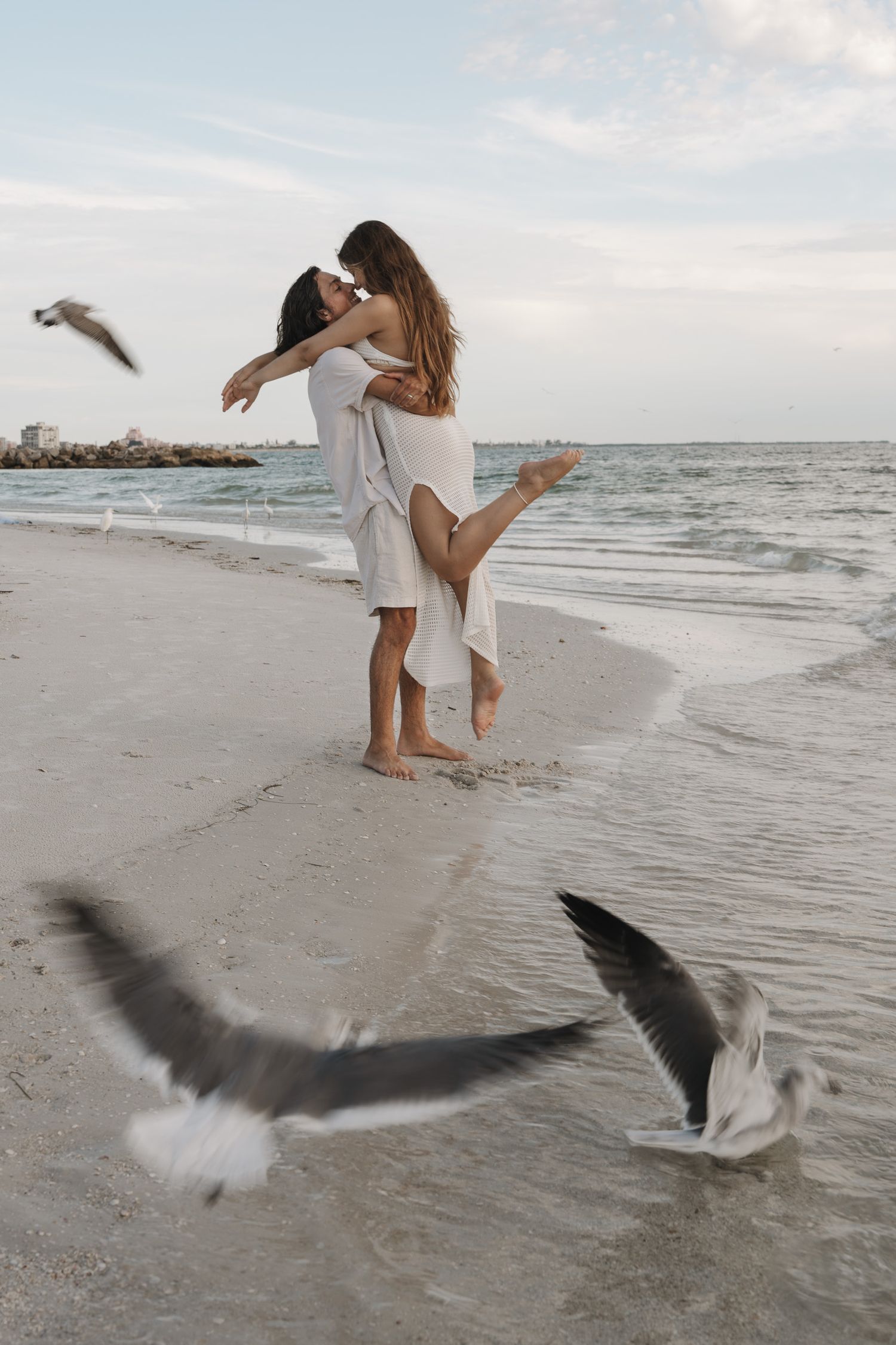A romantic couple at a St. Pete Beach in Florida celebrating their engagement at sunset with seagulls flying and waves in the background.
