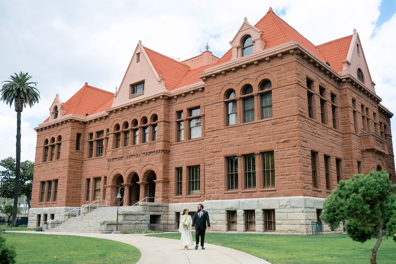 Historic red brick courthouse with Romanesque architecture, arched windows, and red roof, surrounded by green lawn and palm trees.