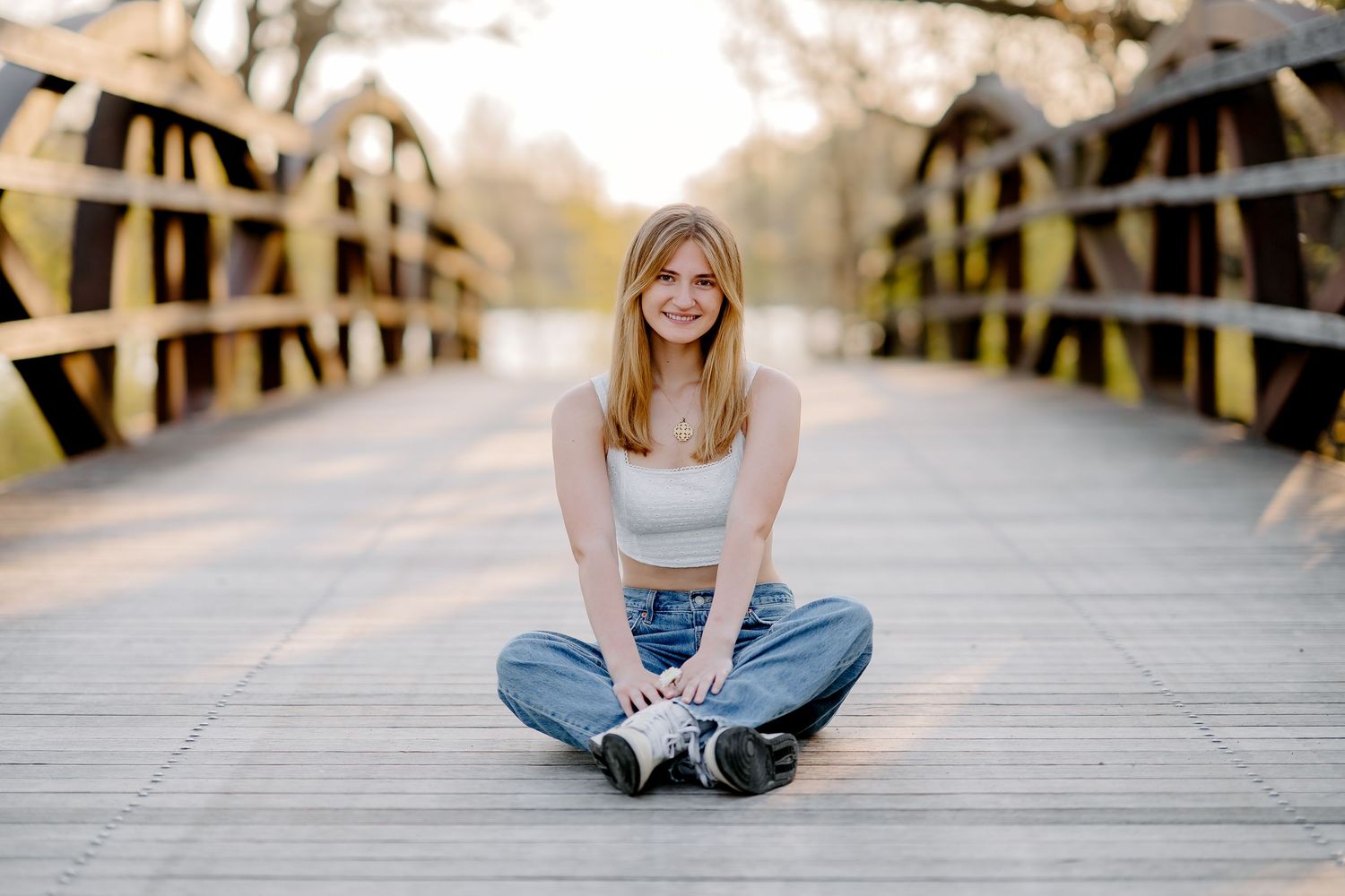 Young blonde woman sitting cross-legged on a wooden bridge, smiling, wearing jeans and a white tank top.