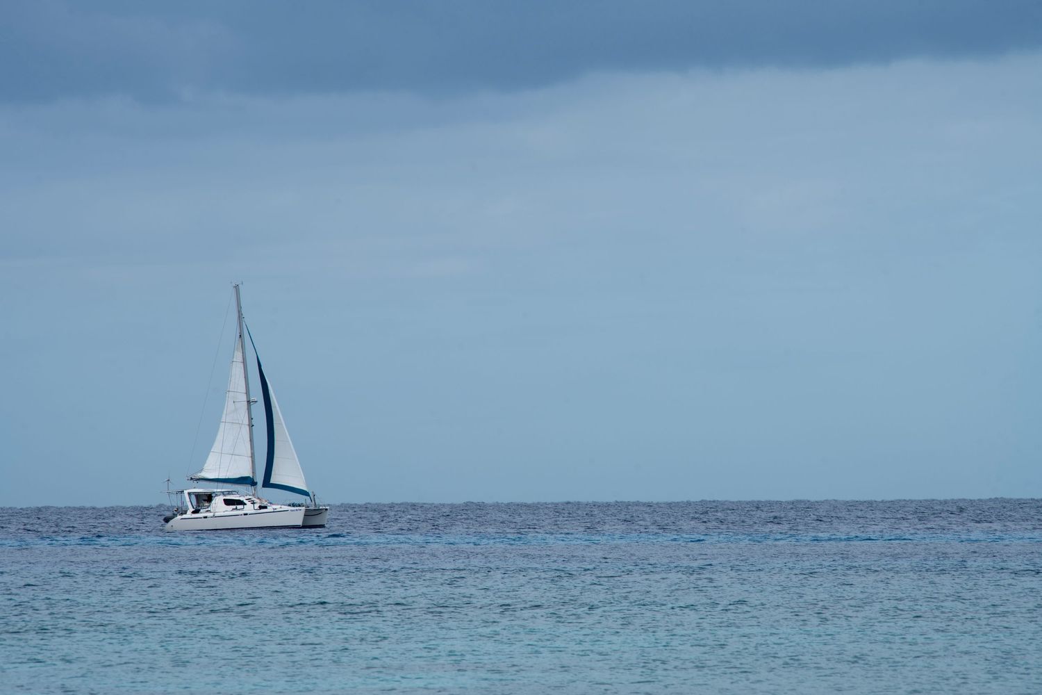White sailboat with blue sails gliding on calm turquoise ocean waters under a cloudy blue sky.