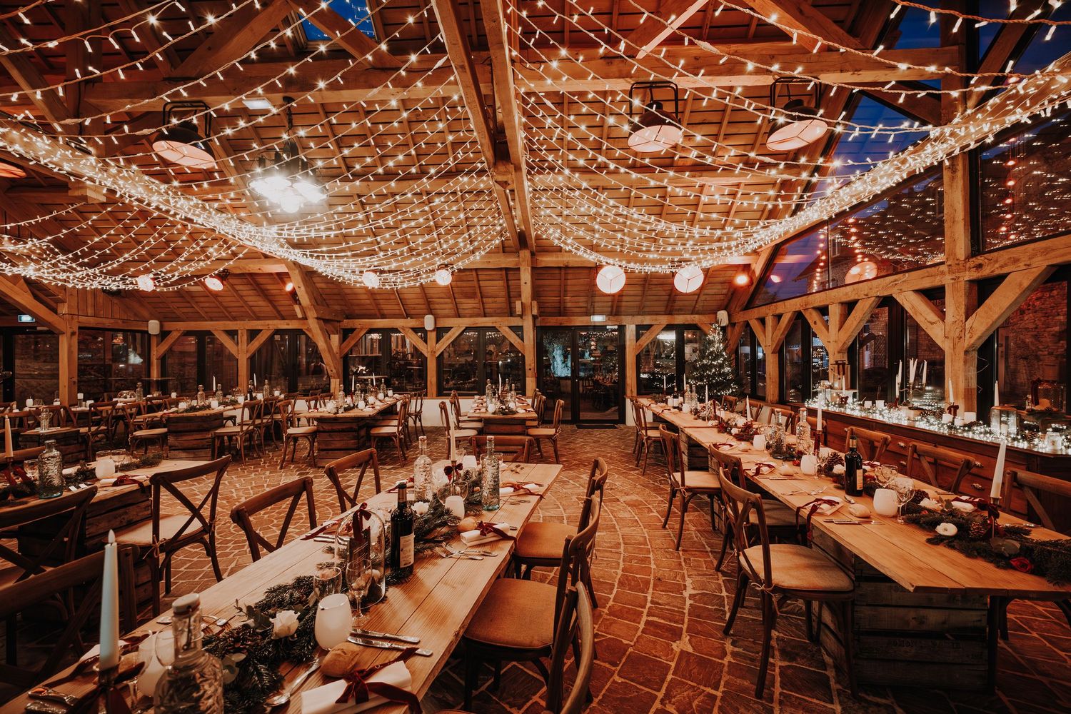 Rustic barn wedding reception with string lights, wooden tables, cross-back chairs, and floral centerpieces on terracotta floors.