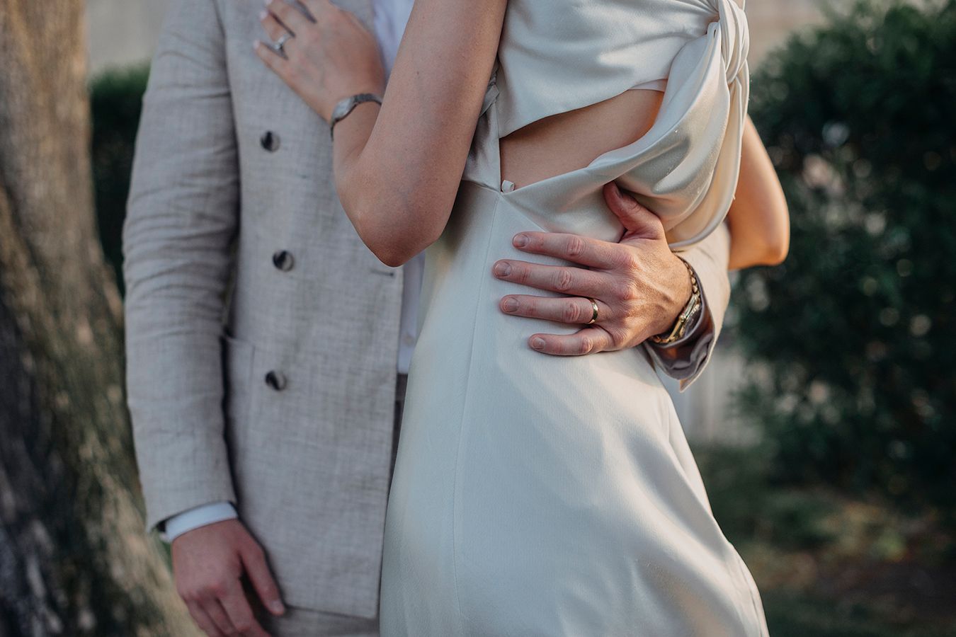 Couple embracing outdoors, woman in white dress and man in beige suit, showing wedding rings and a leather watch.