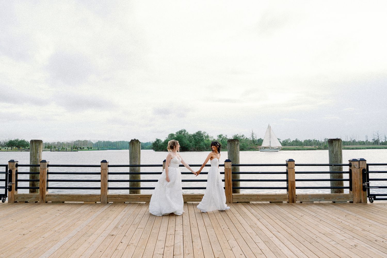 Two brides in white gowns hold hands on a waterfront wooden pier on their wedding day.