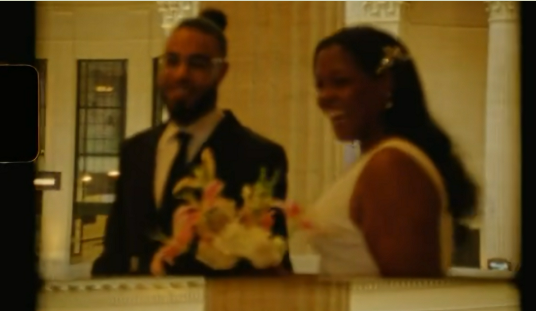 Couple in wedding attire standing at altar, groom in suit with boutonniere, bride in white dress.
