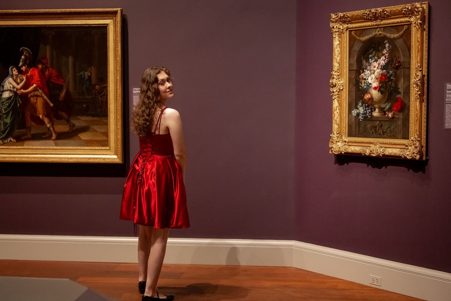A person in a flowing red dress poses dramatically in an art gallery with ornate gold-framed paintings on the walls.