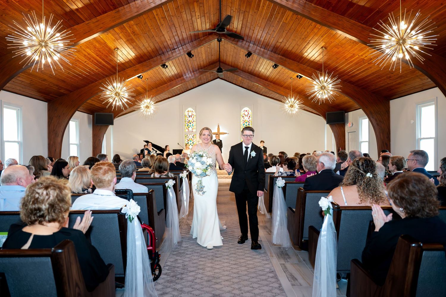 Bride and groom walk down the aisle of a charming chapel with wood-paneled ceiling and starburst chandeliers.