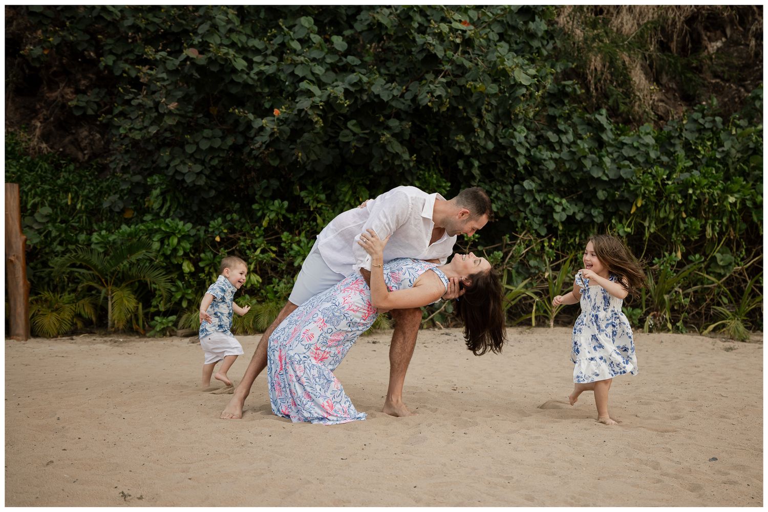 A family plays together on a beach, lifting a child in the air while wearing white clothing in black and white photography.