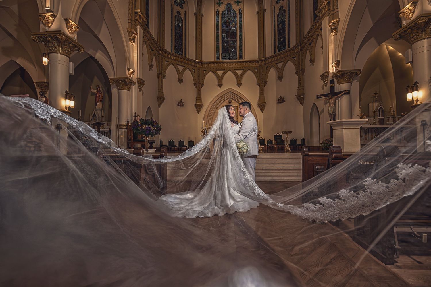 Bride and groom embrace in a majestic cathedral, framed by a long, ornate lace veil sweeping dramatically toward the camera