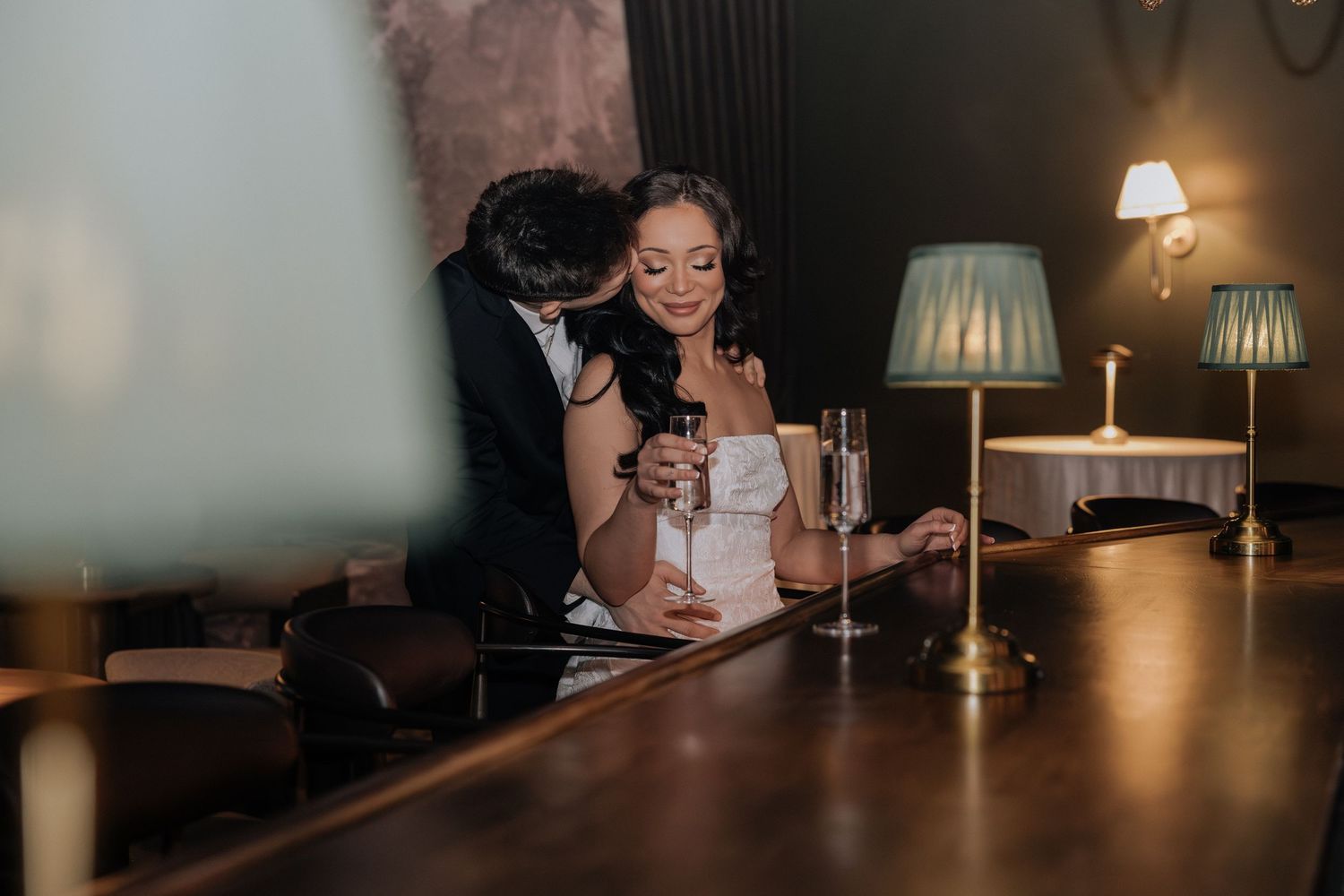 Romantic couple sharing an intimate moment at a dimly lit bar, woman smiling while holding a champagne glass.