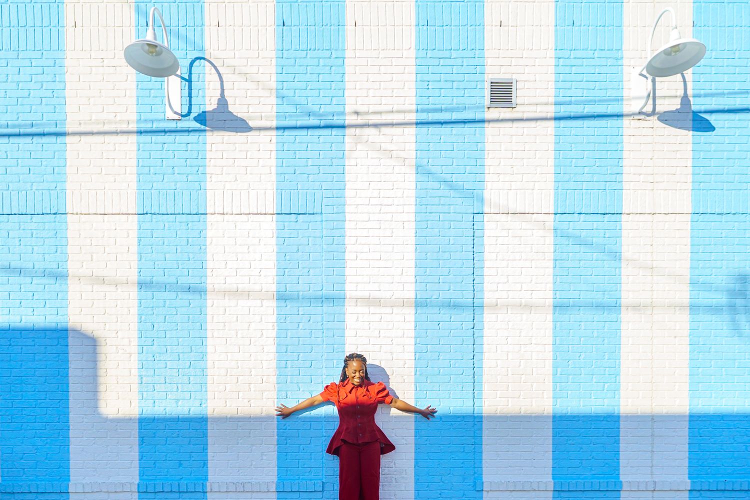 Woman in red dress posing confidently against a bold blue and white striped brick wall outdoors.