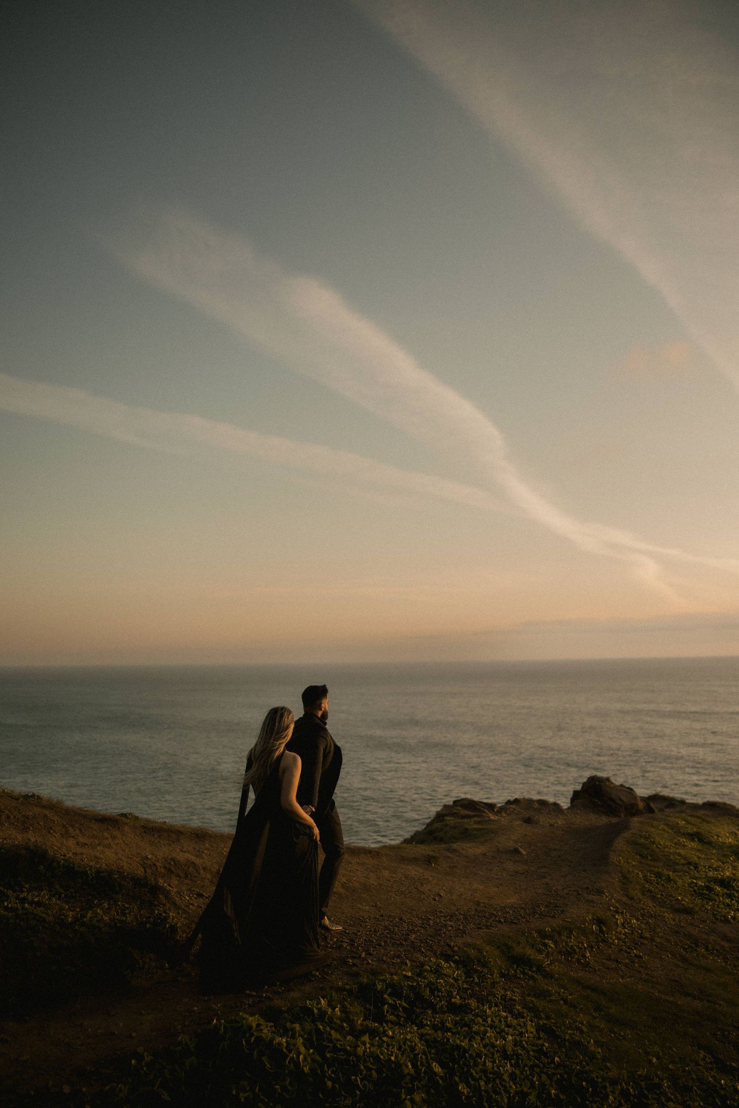 Couple in black attire standing on coastal cliffside at sunset, overlooking calm ocean waters under a pastel sky.