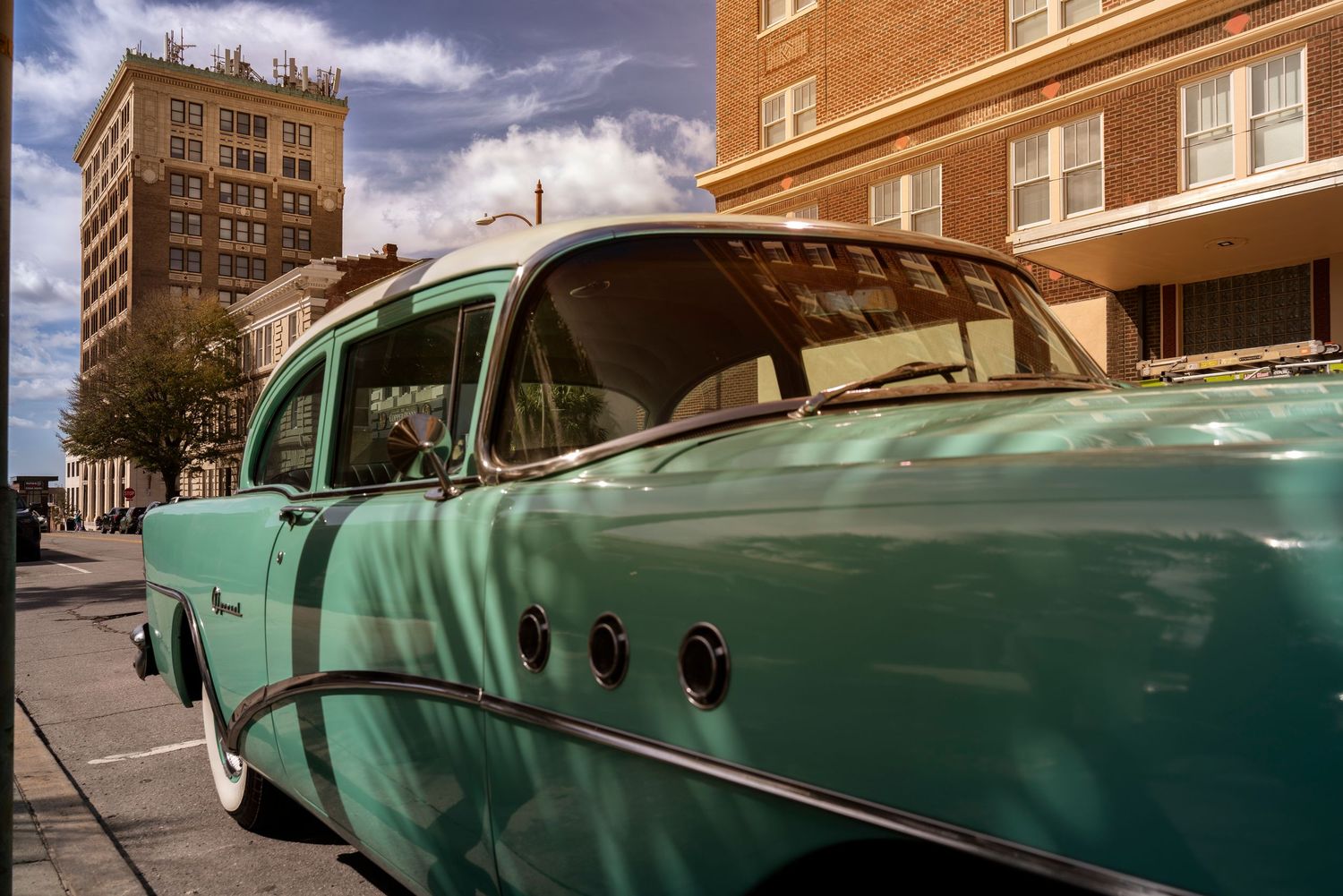 A vintage green car cruises down a city street with tall brick buildings in the background.