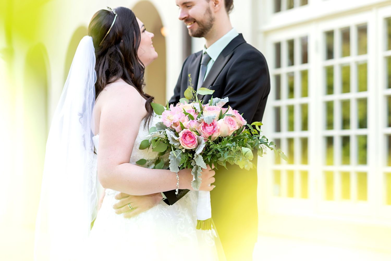 Bride and groom sharing a romantic moment, bride holding a pink rose bouquet, wearing wedding dress with veil outdoors.