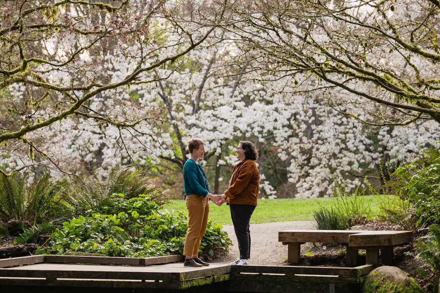 Couple holds hands on a garden path under arching magnolia trees in bloom.