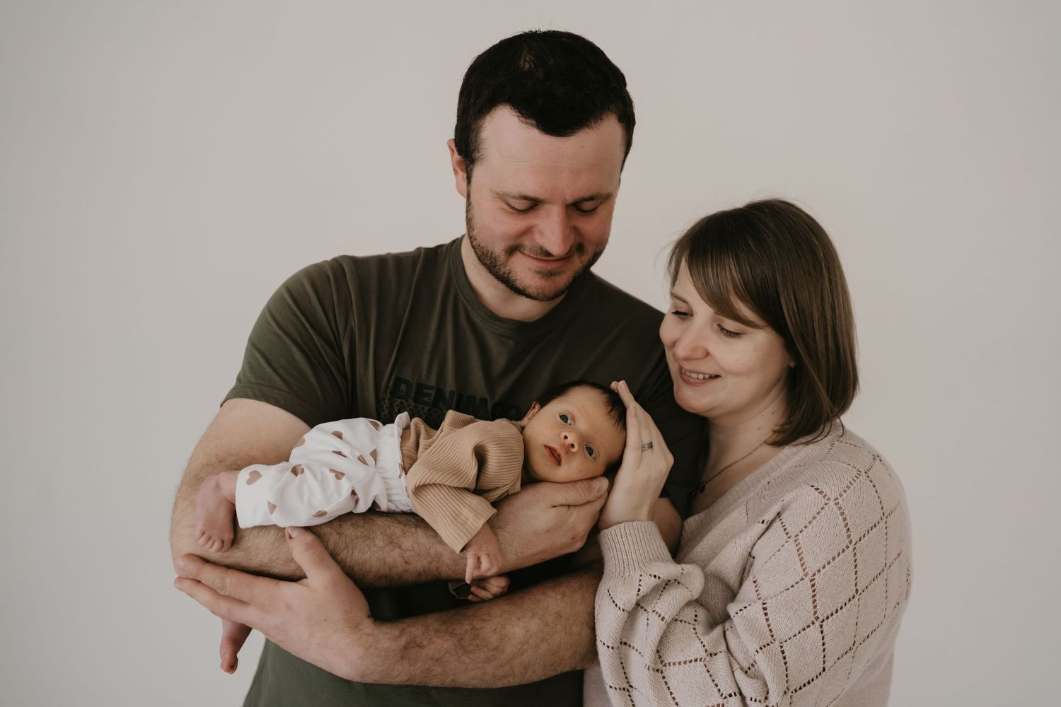 Parents in casual clothing share a tender moment while holding their sleeping newborn baby against a light gray background.