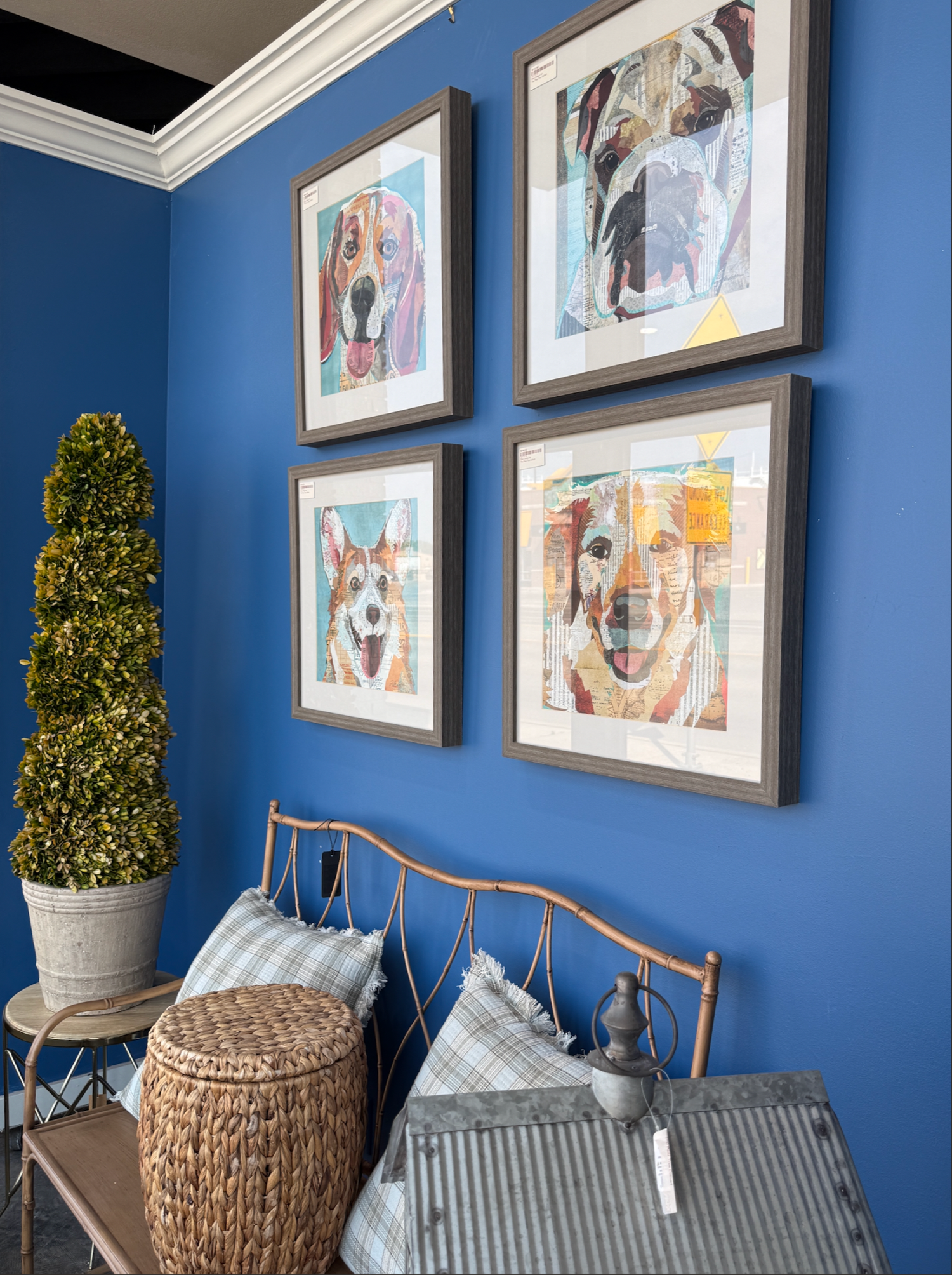 Blue bedroom wall with four framed abstract art prints above a brass bed frame, wicker basket, and decorative topiary plant.