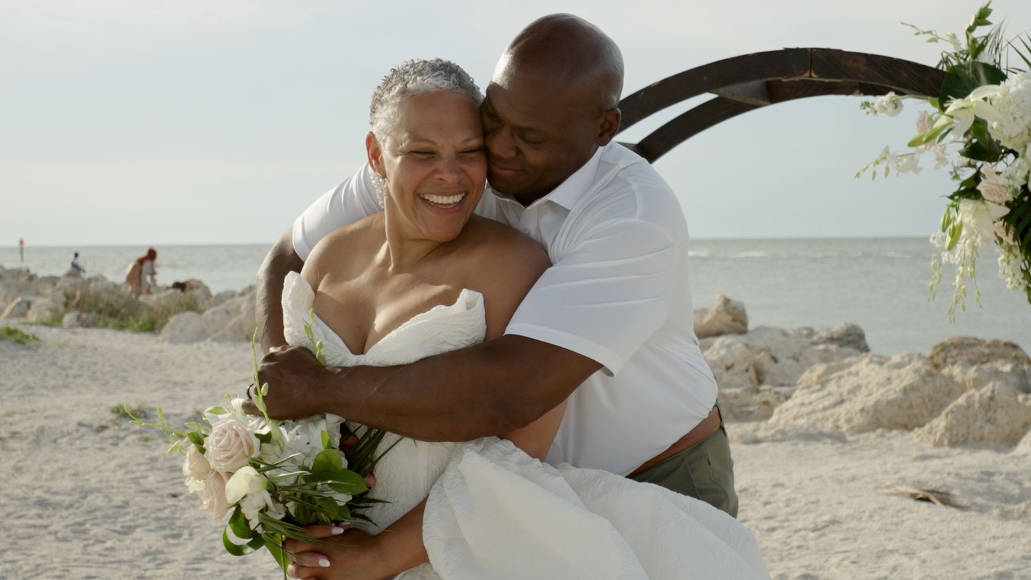 Happy interracial couple embracing on a beach wedding, bride in white dress holding bouquet, groom in white shirt.