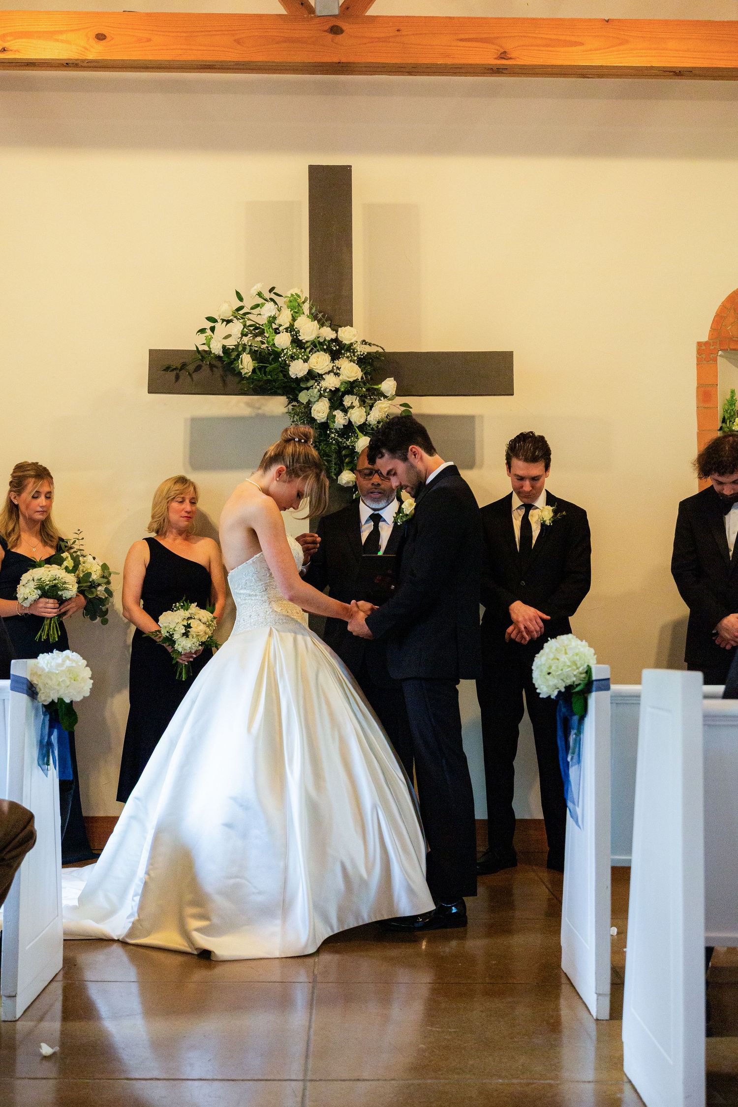 Bride and groom exchange rings at church altar, flanked by bridesmaids in black and groomsmen, with floral cross backdrop.