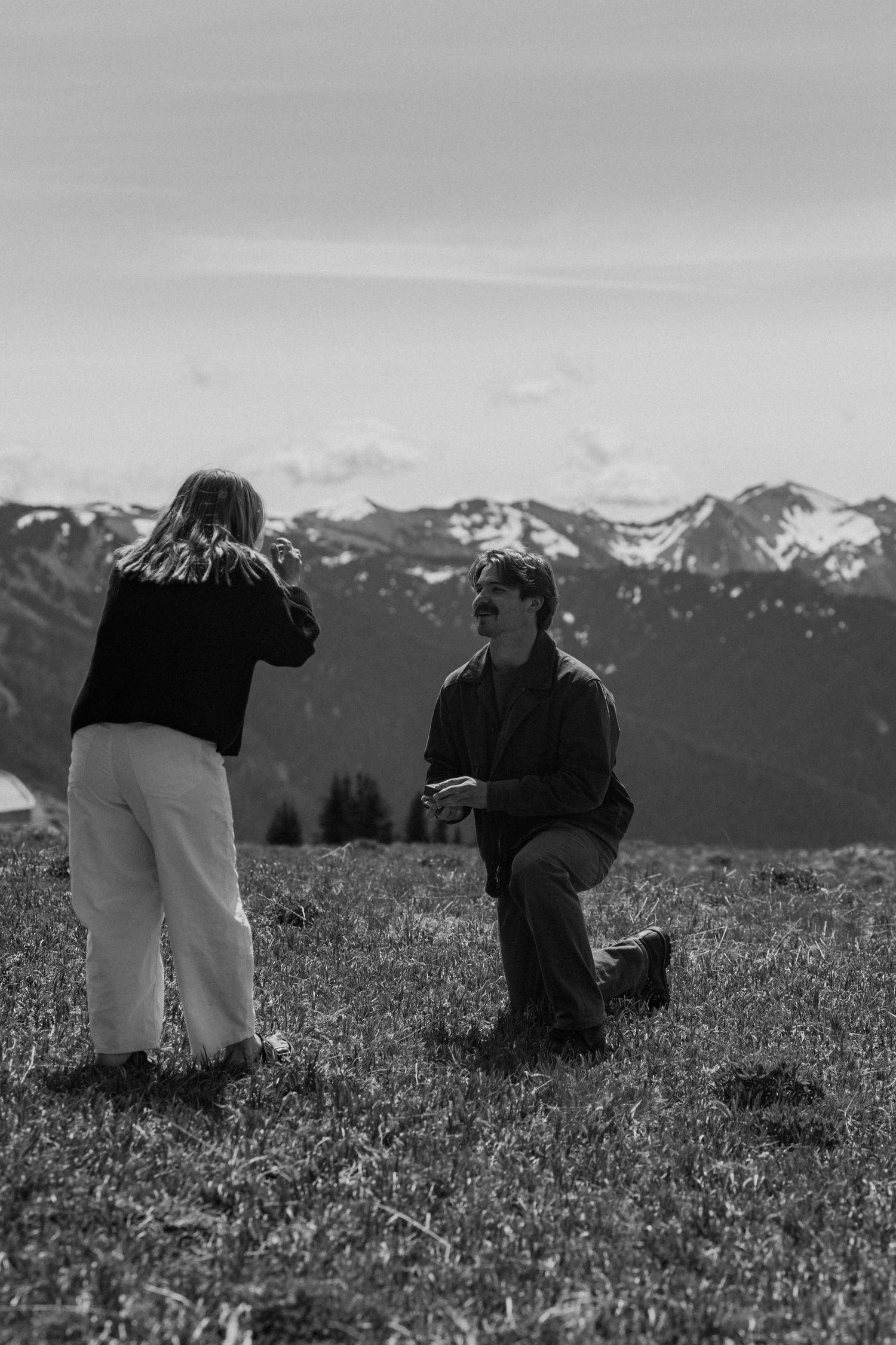 Black and white photo of a man on one knee proposing to a woman on a mountain meadow with snowy peaks in the background.