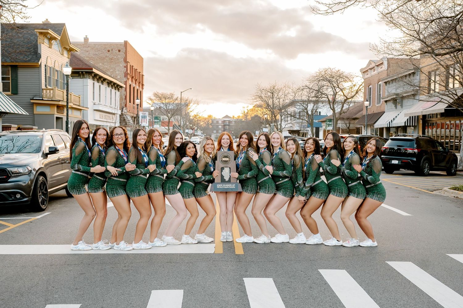 A cheerleading squad in green uniforms poses on a crosswalk in a small town at sunset.