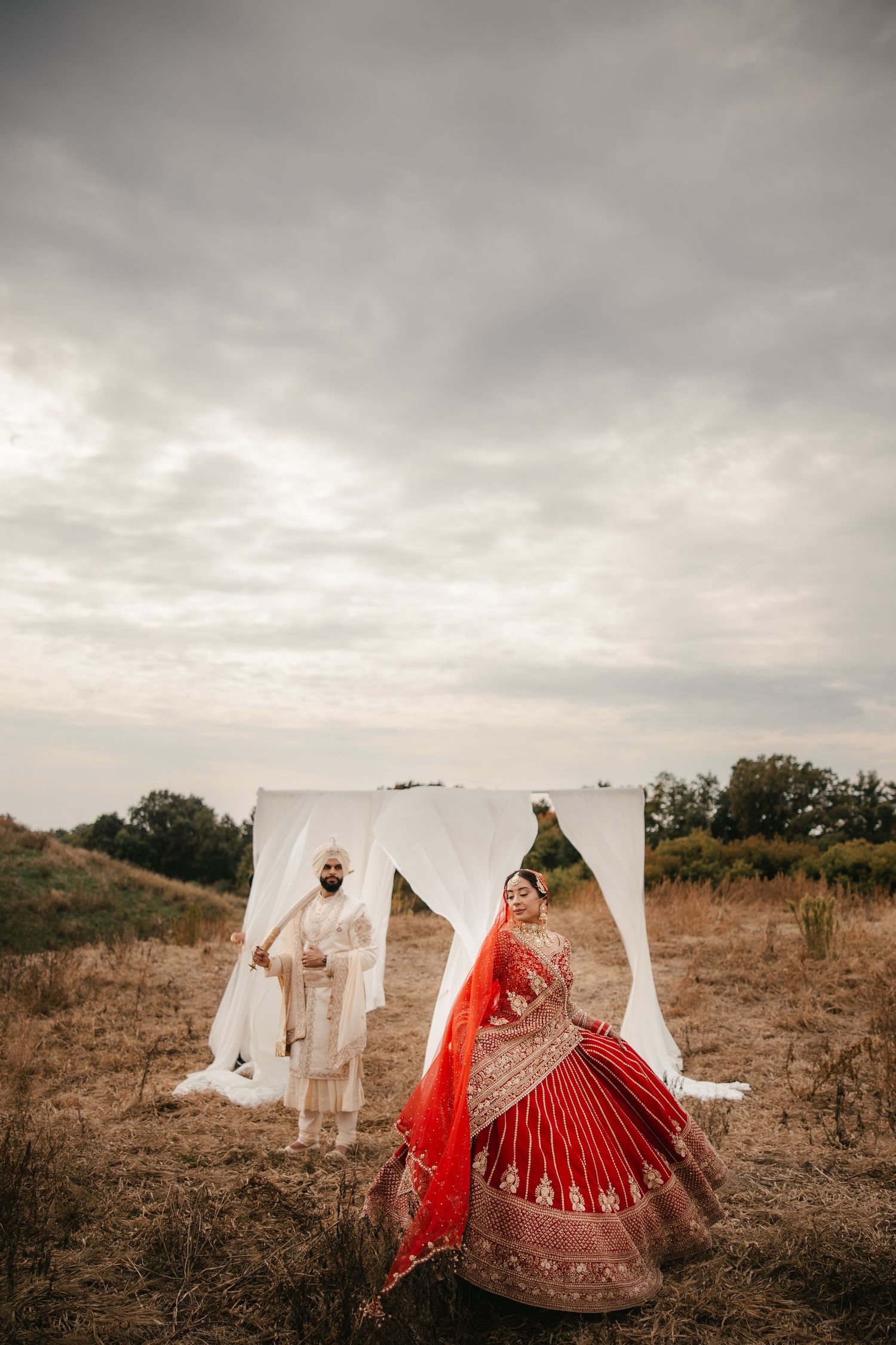 Indian bride in red lehenga and groom in white sherwani share a romantic moment outdoors at their traditional wedding.