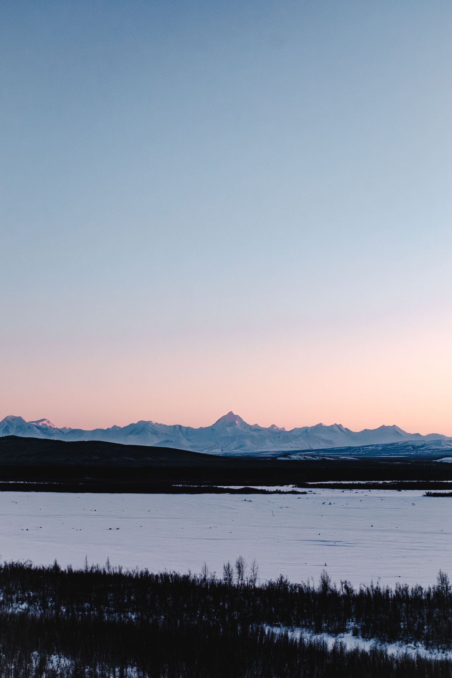 Snow-covered boreal forest valley with frozen river and majestic mountain range under blue sky in winter wilderness.