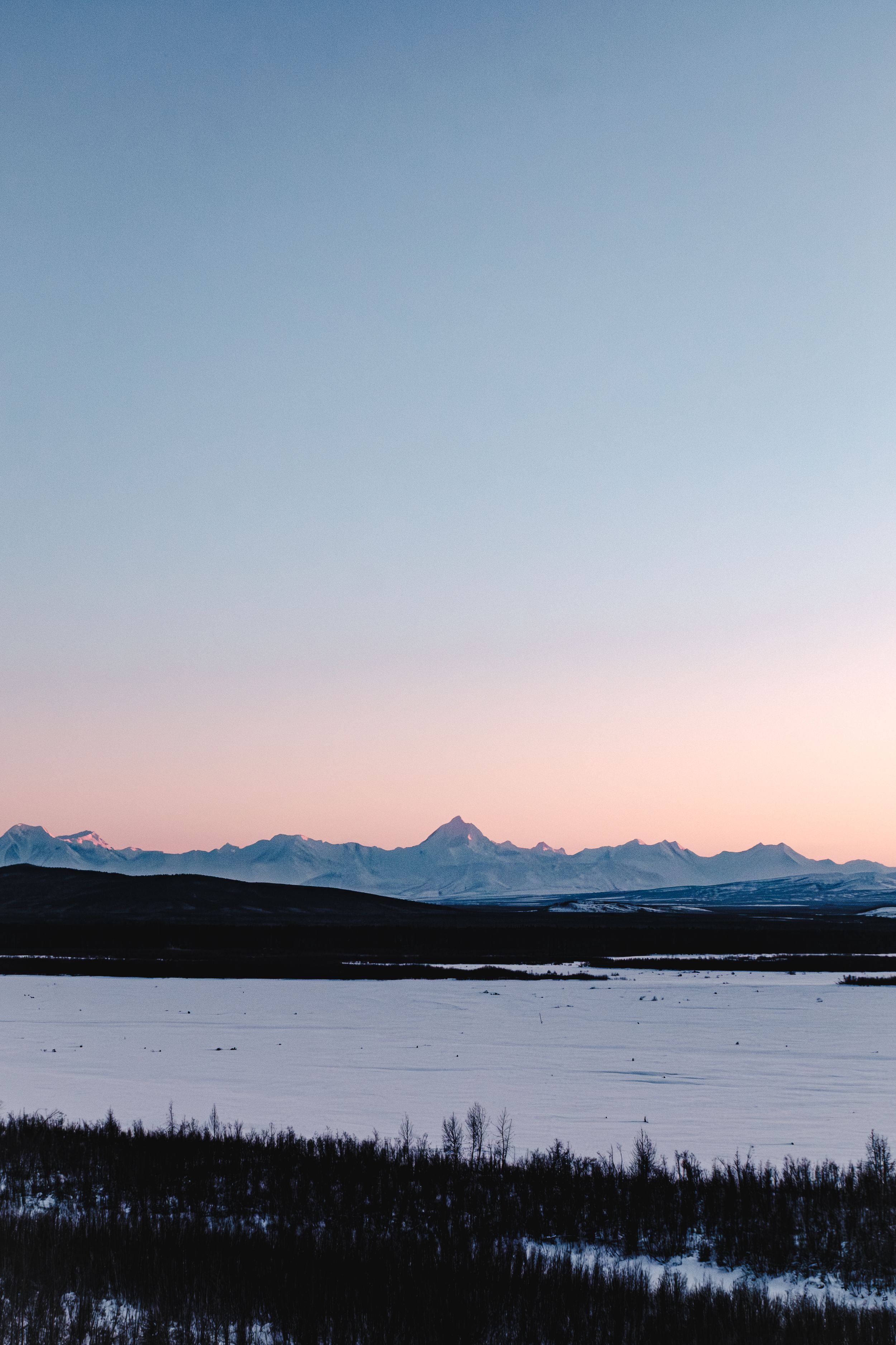 Snow-covered boreal forest valley with frozen river and majestic mountain range under blue sky in winter wilderness.