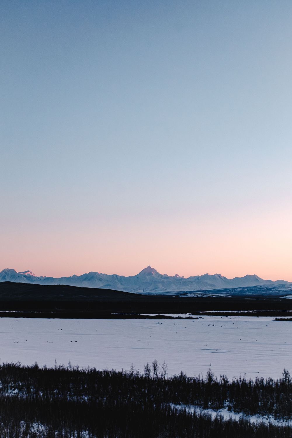 Snow-covered boreal forest valley with frozen river and majestic mountain range under blue sky in winter wilderness.