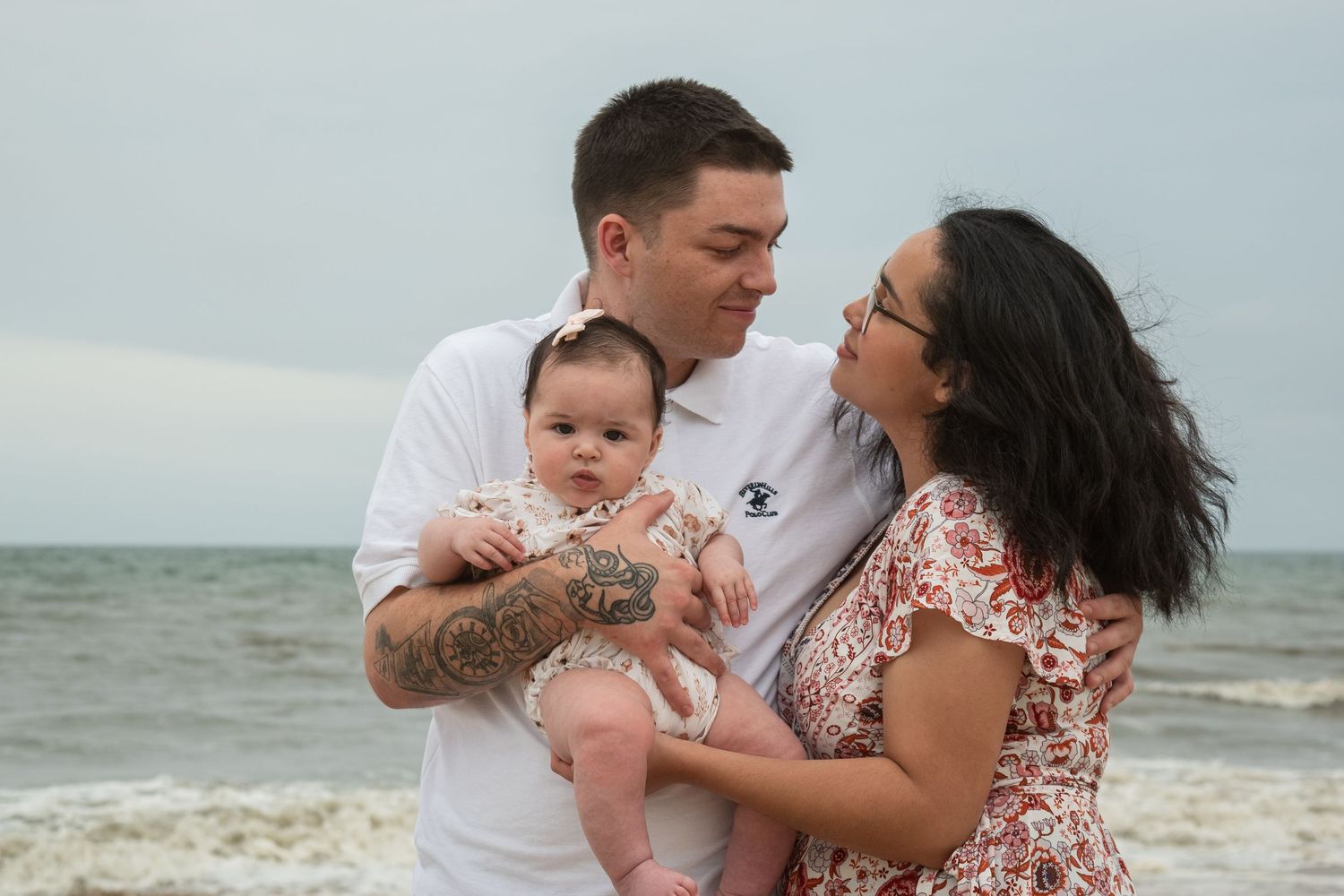 A couple stands on a beach holding their baby, with ocean waves in the background on a cloudy day.