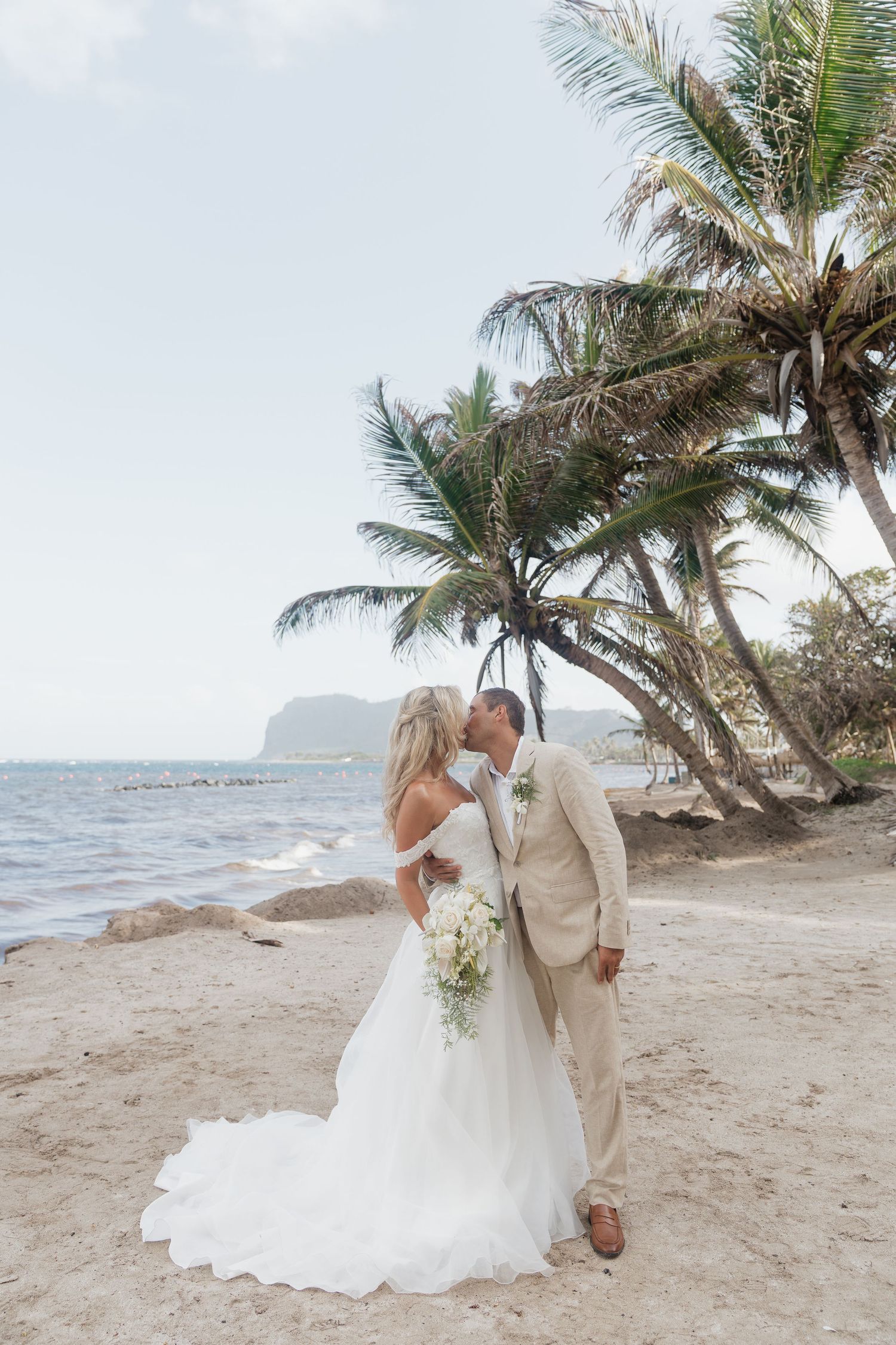 Bride and groom kissing on a St Lucia tropical beach with palm trees, bride in white gown holding bouquet, groom in tan suit.