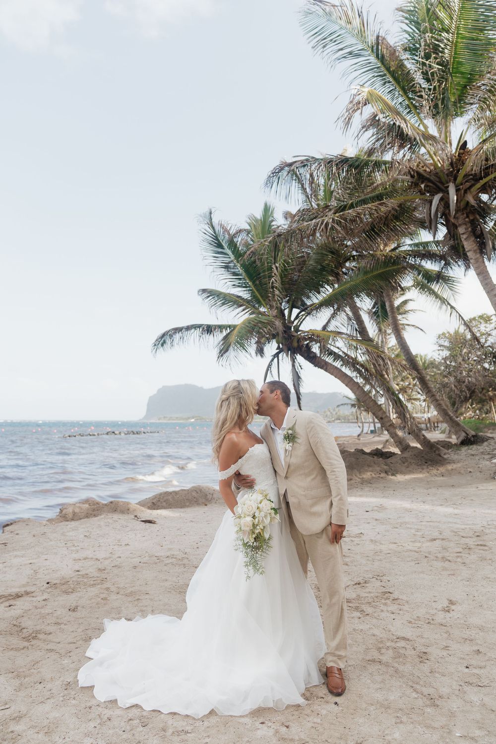 Bride and groom kissing on a St Lucia tropical beach with palm trees, bride in white gown holding bouquet, groom in tan suit.