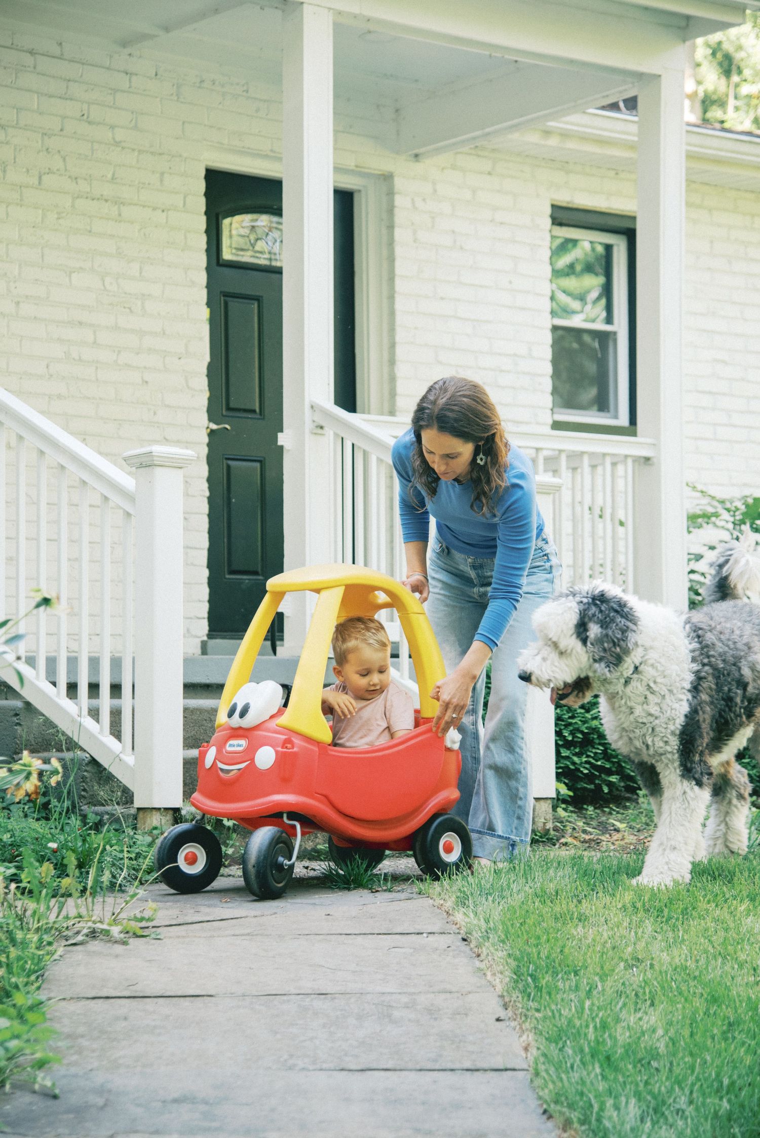 Family with toddler and Old English Sheepdog playing on white brick home front porch steps on sunny day.