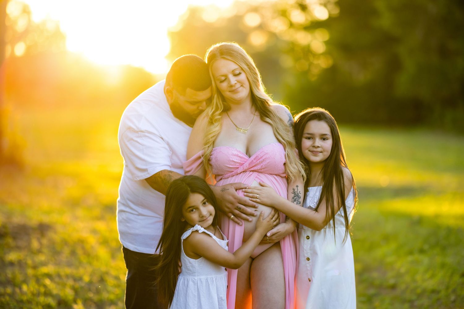Pregnant woman in pink dress poses with family at golden hour in a sunny outdoor maternity photo session.