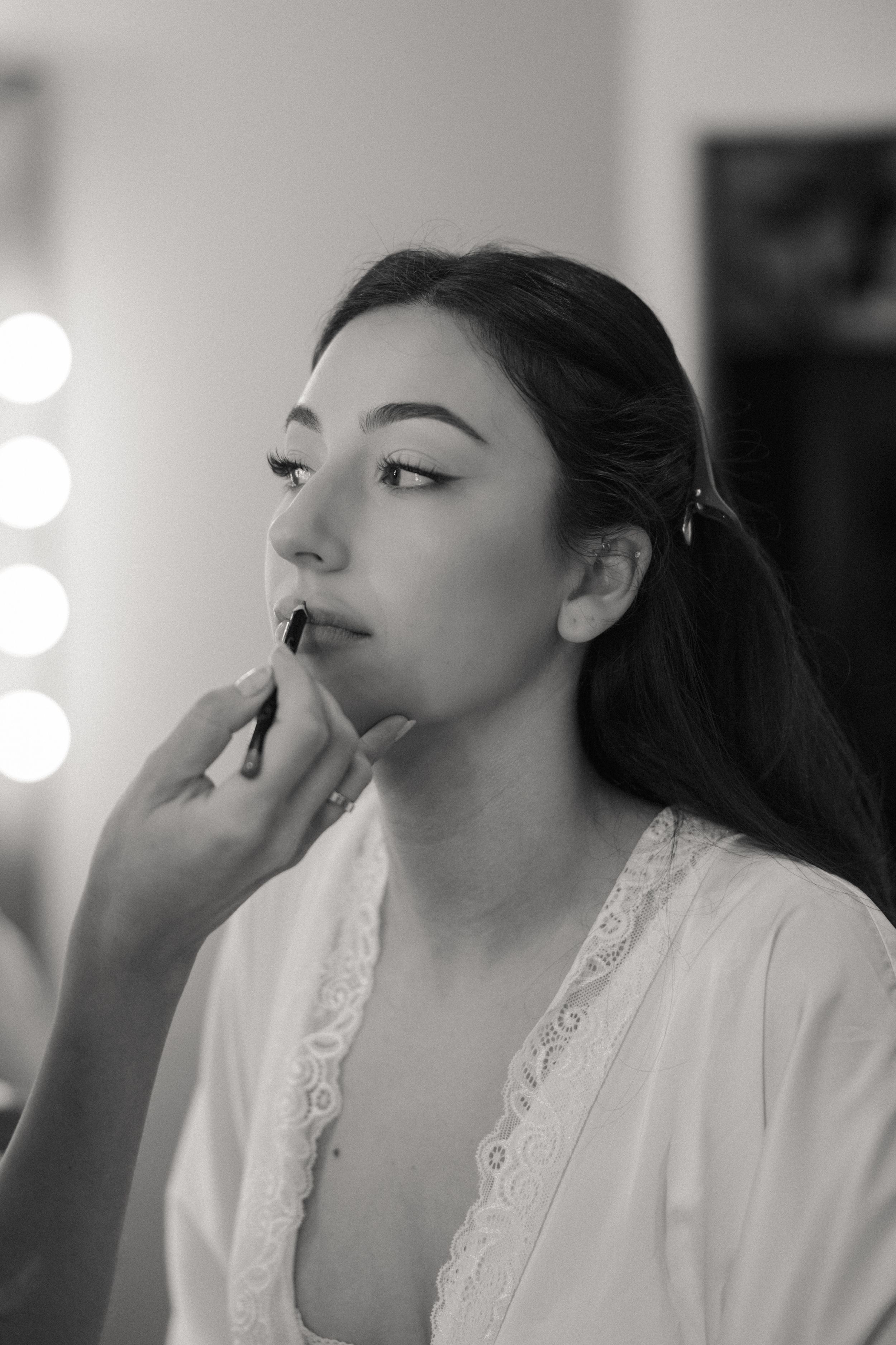 Black and white photo of a young woman getting lipstick applied by a makeup artist in a vanity mirror setting.