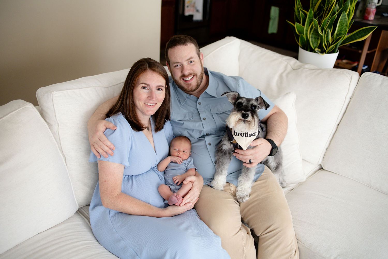 Smiling couple sits on white sofa with newborn baby and schnauzer dog indoors.