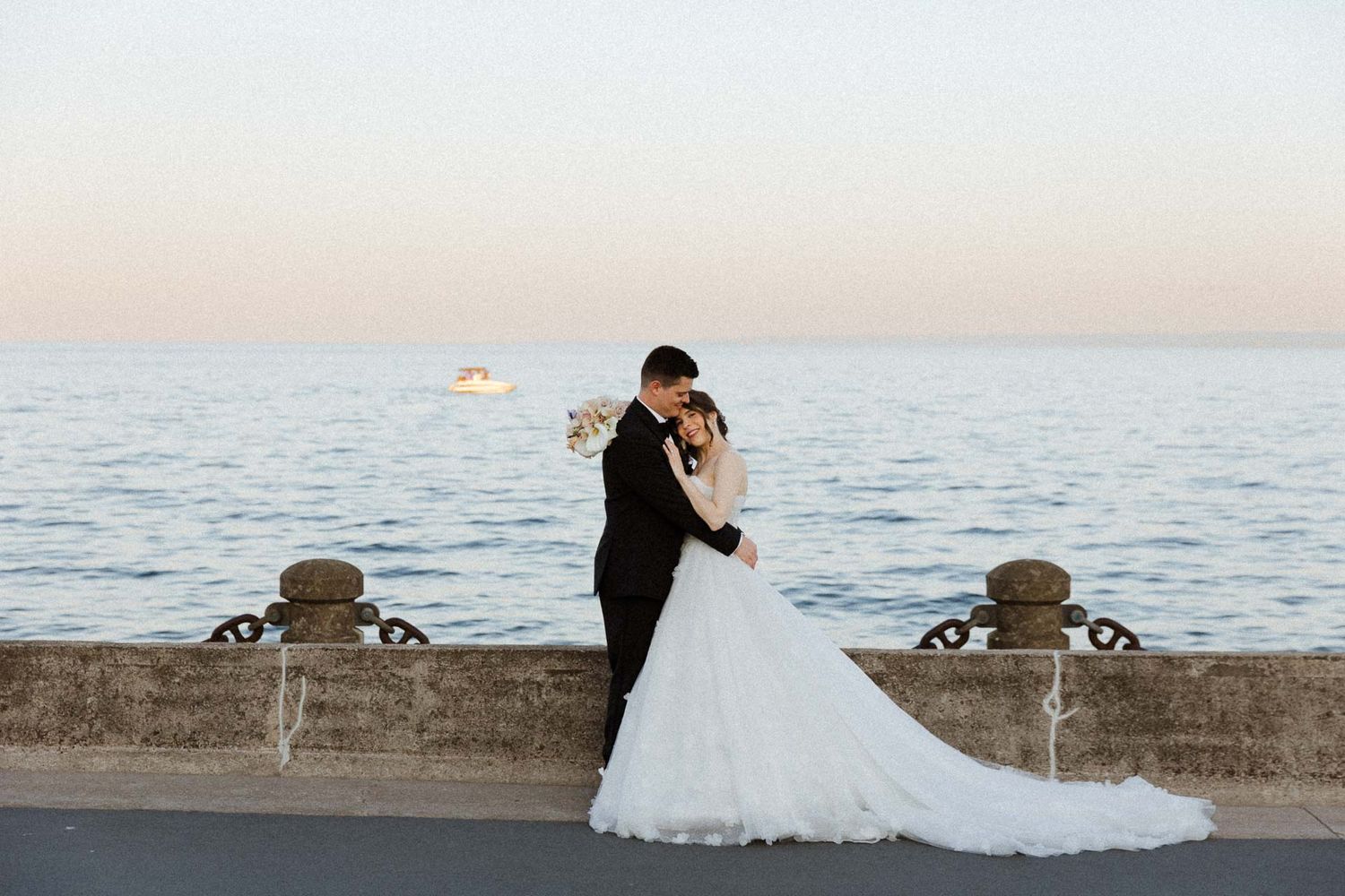 A couple in wedding attire embraces at sunset by an ornate stone wall overlooking the ocean.