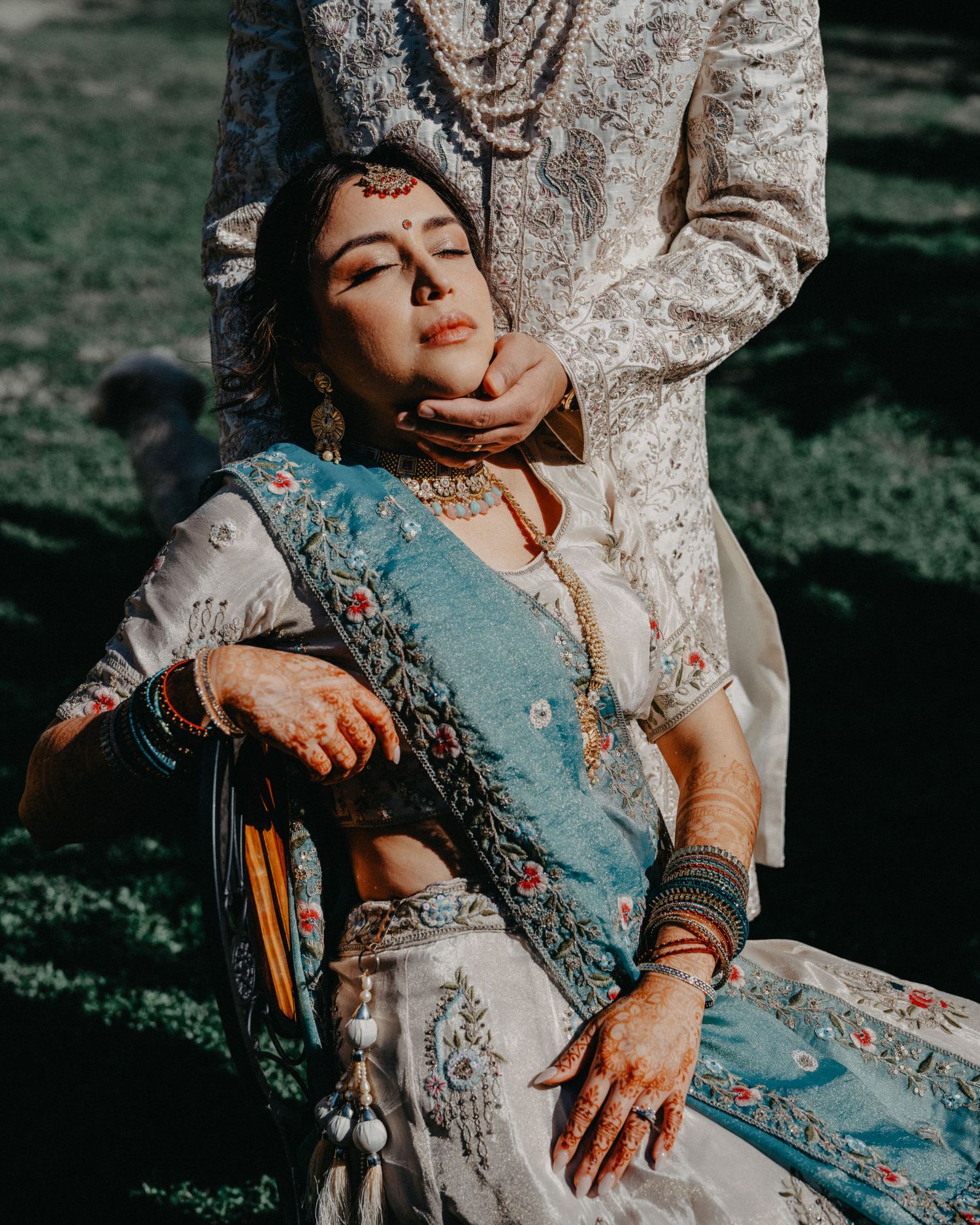 A person in an ornate light blue traditional dress with embellishments poses dramatically in sunlit outdoor setting.