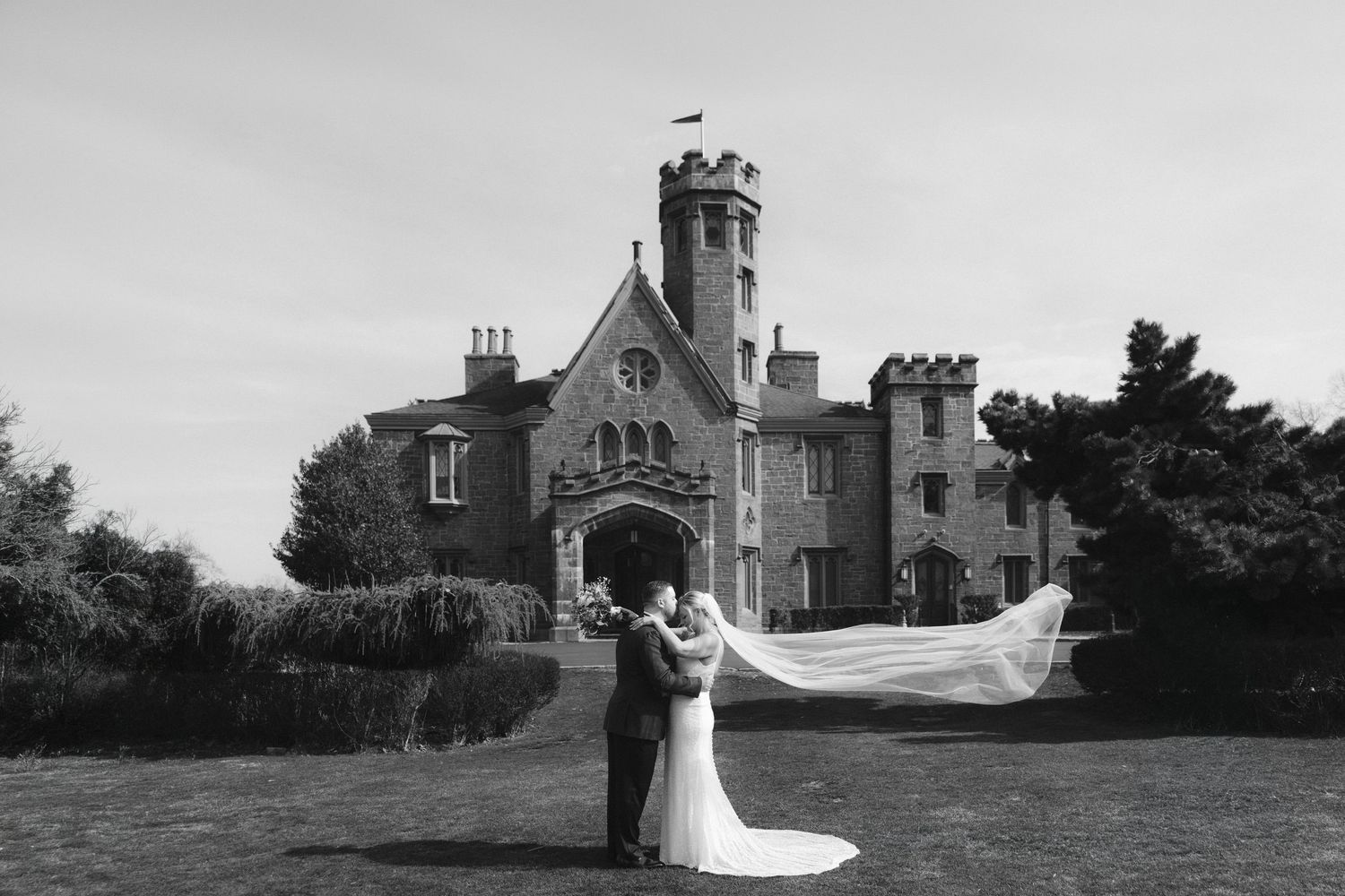 Outdoor wedding ceremony takes place on lawn in front of grand stone castle with Gothic architecture under cloudy skies.