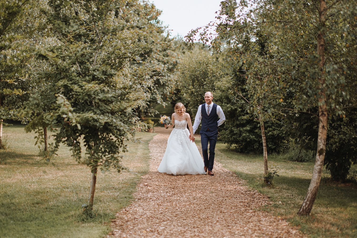 Newlyweds walking hand in hand down a tree-lined garden path.