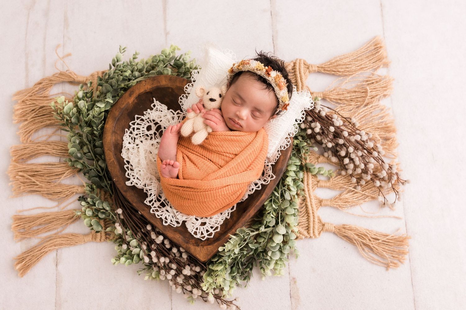 Sleeping newborn swaddled in orange wrap nestled in a heart-shaped wooden bowl with floral greenery decor.