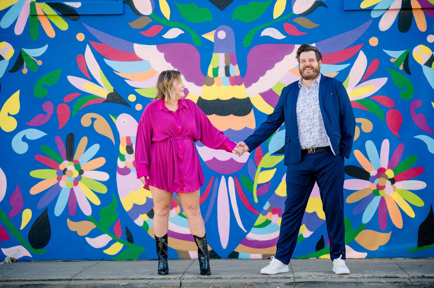 Couple posing playfully against vibrant floral mural wall art in bright colors.