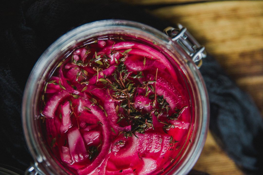 Glass jar of vibrant pink pickled vegetables with fresh herbs sprinkled on top.