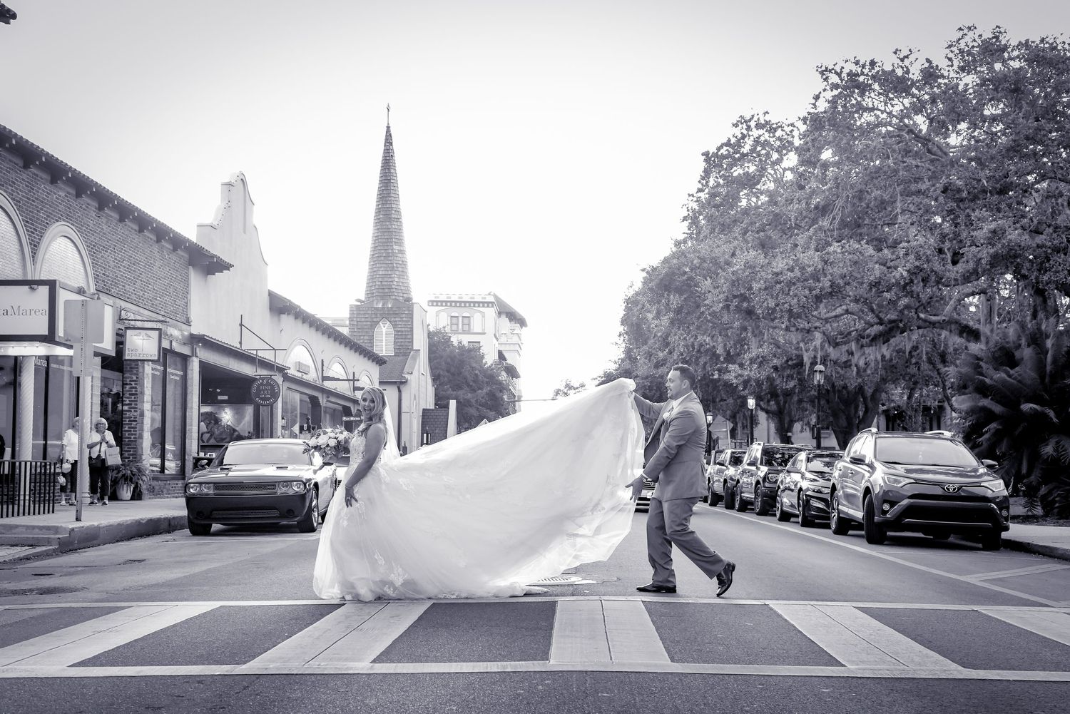 A dramatic black and white photo of a flowing wedding dress on a street with a church spire in the background.