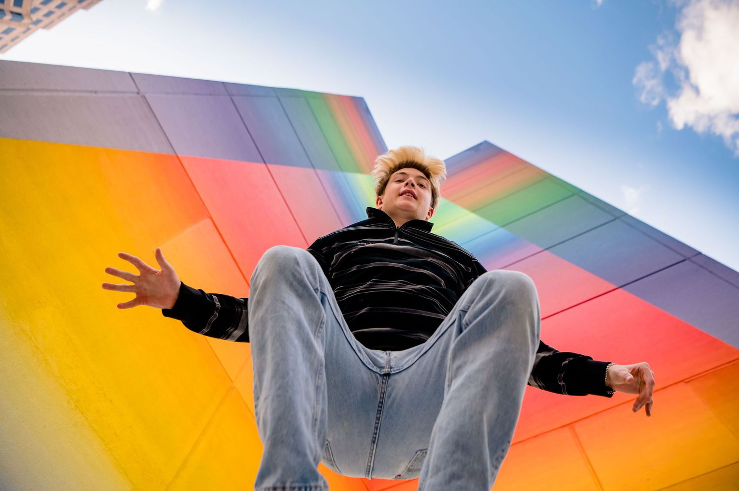 Person jumping joyfully against a colorful geometric rainbow background outdoors.