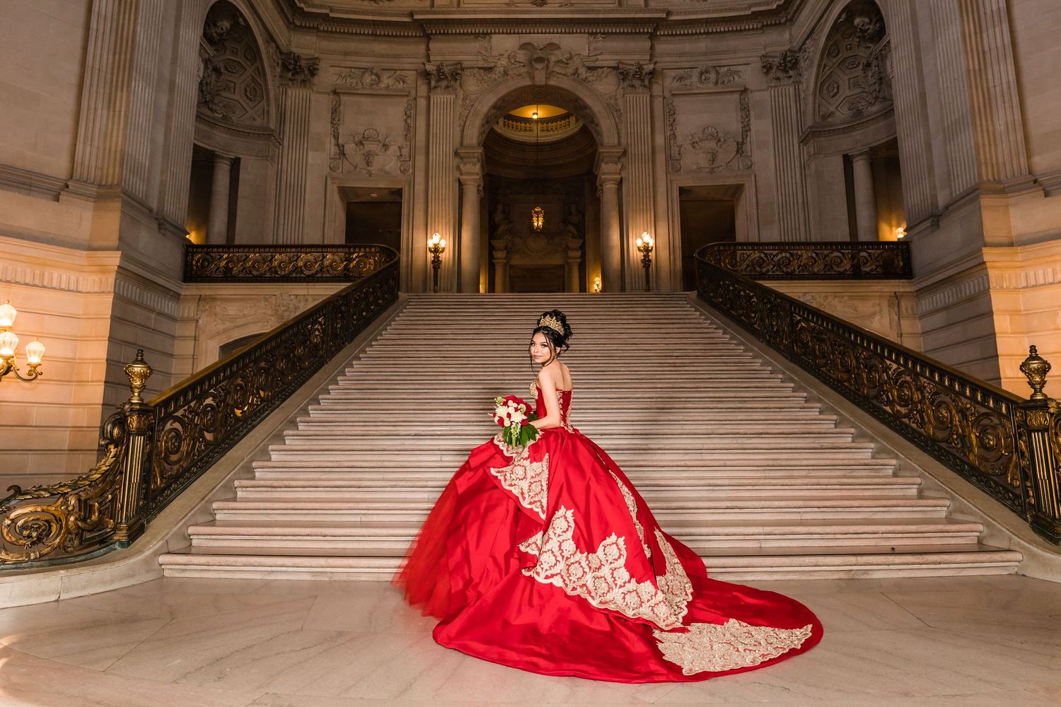 An elegant red ball gown with gold accents photographed on grand marble steps with ornate railings.